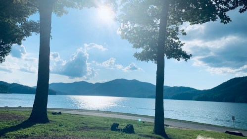 A serene lake view framed by two trees, with mountains in the background and sunlight reflecting on the water.