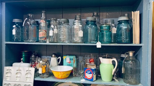 A wooden shelf filled with various glass jars, bottles, and kitchenware, some with price tags attached.