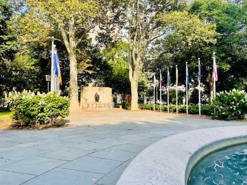 A peaceful park scene featuring a fountain, flags, and a memorial surrounded by trees and greenery.