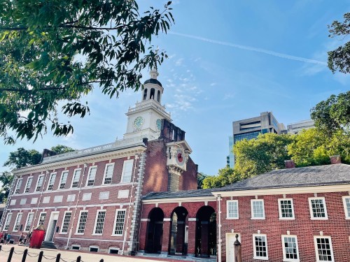 Historic building with a clock tower, surrounded by trees and modern buildings under a clear blue sky.