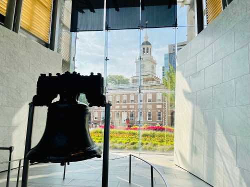 A large bell in the foreground with a view of a historic building and colorful flowers through glass walls.