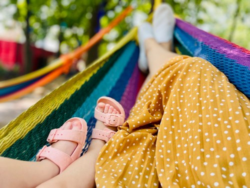 A person in a yellow polka dot dress relaxes in a colorful hammock, with a child's feet in pink sandals nearby.
