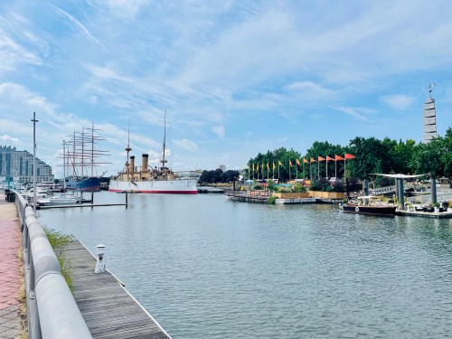 A scenic harbor view featuring historic ships, colorful flags, and lush greenery under a blue sky.