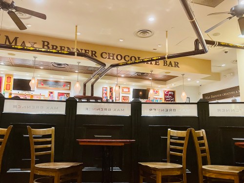 Interior of Max Brenner Chocolate Bar, featuring wooden chairs and a counter with colorful decor in the background.