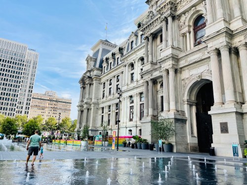 A person stands near a fountain in front of a grand historic building on a sunny day.