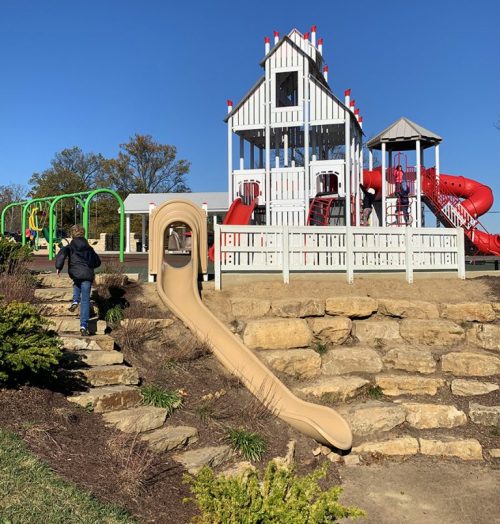 A playground with a large play structure, slide, and climbing equipment, surrounded by greenery and stone steps.