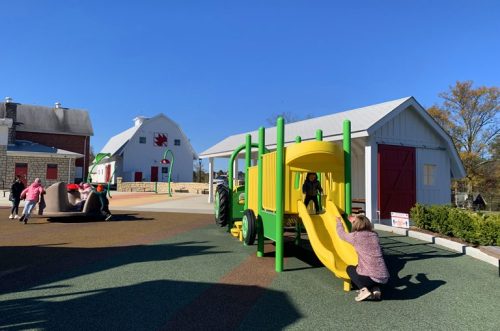 A colorful playground with a yellow slide, children playing, and a barn in the background under a clear blue sky.