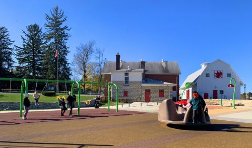 A playground with swings and a spinning structure, set against a clear blue sky and a historic building in the background.