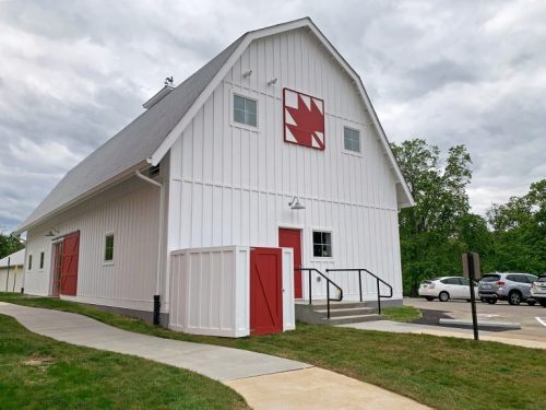 A white barn with a red emblem and doors, surrounded by green grass and a paved walkway, under a cloudy sky.