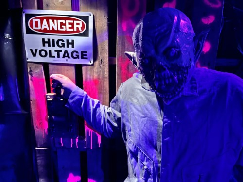 A creepy figure with a monstrous face stands next to a "Danger High Voltage" sign in a dimly lit setting.