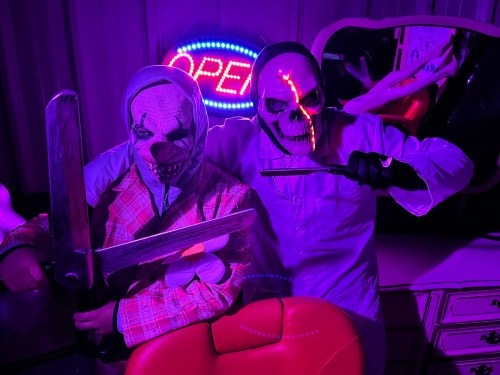 Two figures in creepy costumes hold large blades, surrounded by purple lighting and an "OPEN" neon sign.