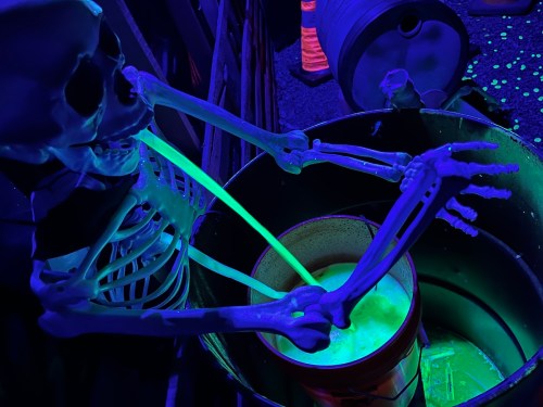 A skeleton with glowing green arms leans over a bucket, surrounded by colorful lights and a dark background.