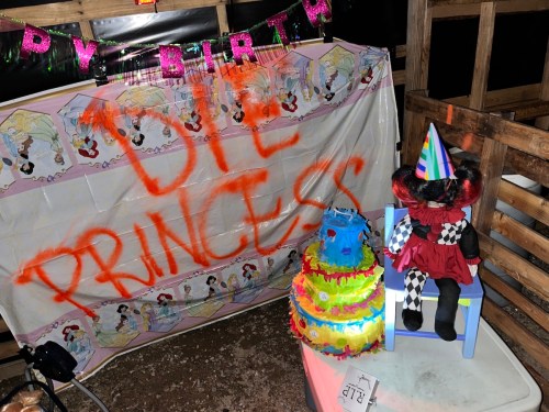 A birthday setup with a colorful cake, a doll in a party hat, and a banner with "Happy Birthday" and "Die Princess" written on it.