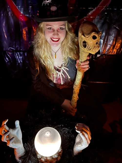 A person in a top hat holds a skull-topped staff, sitting near a glowing crystal ball and skeletal hands.