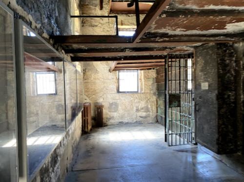 Interior of a stone-walled room with a barred door, windows, and exposed wooden beams.