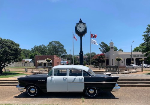 A classic black and white car parked near a clock tower and flags in a sunny town square.