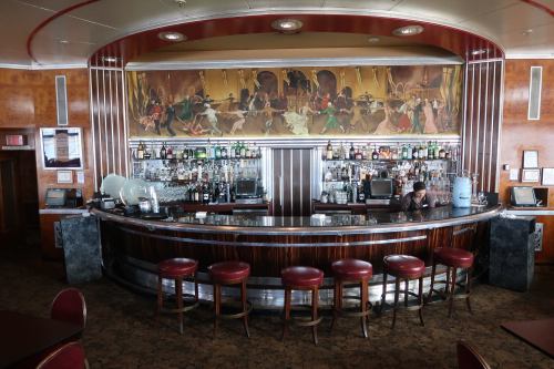 A vintage bar with a curved counter, shelves of liquor, and a mural depicting lively scenes above. Red stools line the bar.