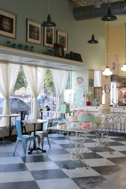 A cozy café interior with checkered floors, tables, and large windows draped with sheer curtains.