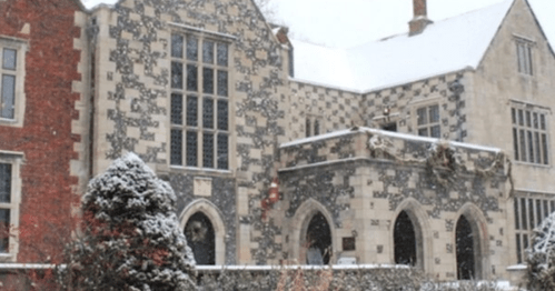A snow-covered historic building with intricate stonework and large windows, surrounded by trees and a winter landscape.