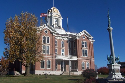 Historic brick courthouse with a clock tower, flag, and a statue in front, surrounded by trees and clear blue sky.