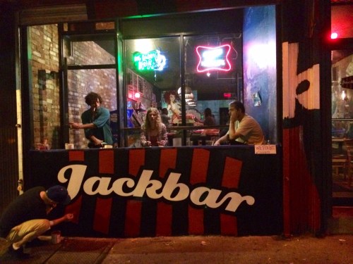 A vibrant bar exterior at night, with people sitting at a painted counter and neon lights glowing in the background.