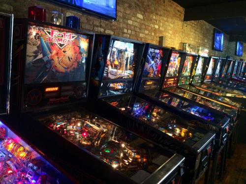 A row of colorful pinball machines in a dimly lit arcade with brick walls and screens in the background.
