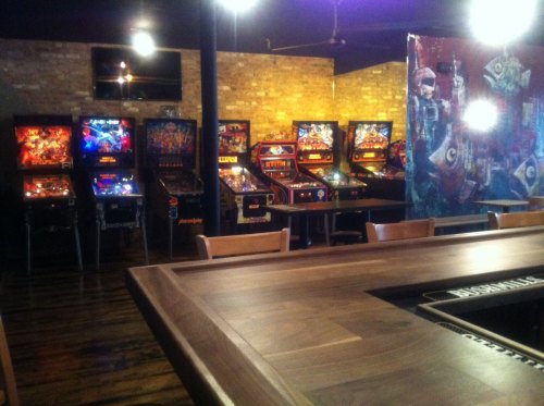 A cozy arcade with several vintage pinball machines lined up against a brick wall, and a wooden bar in the foreground.
