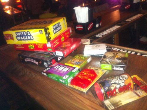 A stack of various board games and card games on a wooden table in a dimly lit setting.