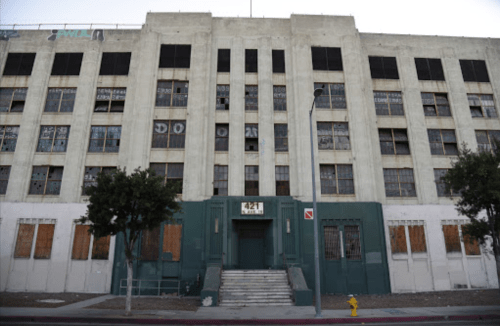 Abandoned building with boarded windows and a worn facade, featuring a central entrance and overgrown surroundings.
