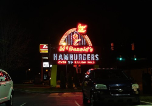 A brightly lit McDonald's sign at night, advertising "Over 99 Billion Sold" with cars parked nearby.