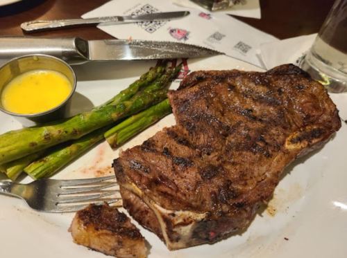 A plate with a grilled steak, asparagus, and a small cup of yellow sauce, with utensils beside it.