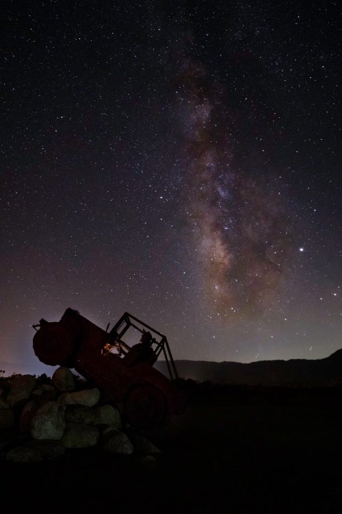 A rusted tractor silhouette against a starry night sky, featuring the Milky Way and distant mountains.