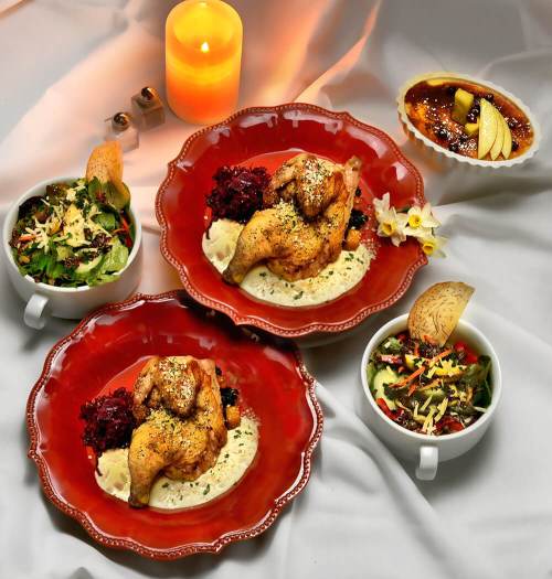 Two plates of roasted chicken with mashed potatoes and salads, accompanied by a dessert, all elegantly arranged.