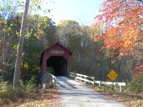 A red covered bridge surrounded by autumn trees, with a sign indicating it's a one-lane bridge.