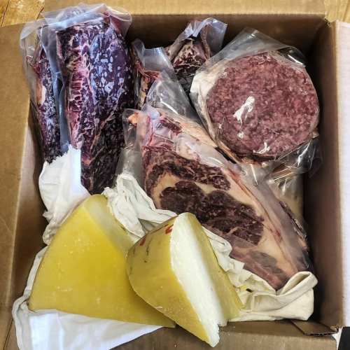 A cardboard box containing various cuts of meat and a block of cheese, all vacuum-sealed in plastic.