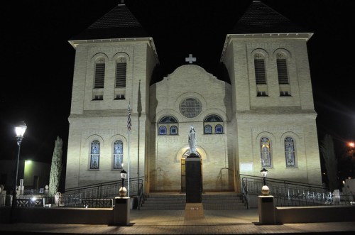 Historic church illuminated at night, featuring twin towers, stained glass windows, and a statue in front.