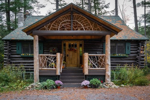 A charming log cabin with a decorative porch, surrounded by trees and colorful flowers in a serene setting.