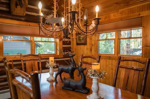 Cozy log cabin dining area with a wooden table, candle holders, and a chandelier above. Rustic decor and natural light.