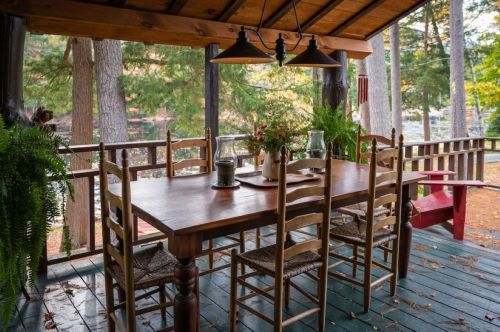 A rustic wooden dining table with chairs on a porch, surrounded by trees and a view of a lake.