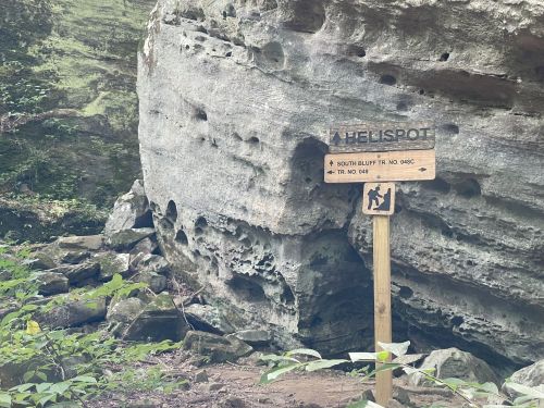 A wooden sign labeled "Heli Spot" near a large rock formation in a forested area, with hiking trail information.