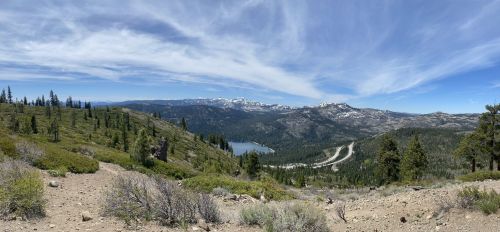 Panoramic view of a mountainous landscape with a lake, trees, and winding road under a blue sky with clouds.