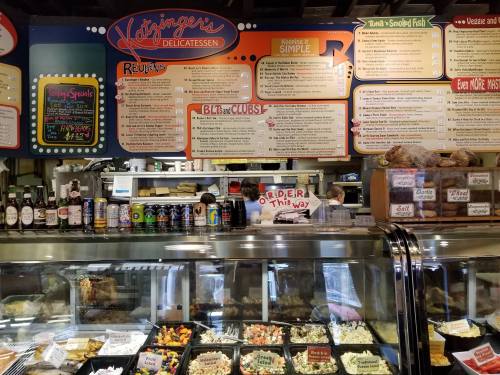 A deli counter with a menu board overhead, showcasing various food items and drinks in a vibrant, busy setting.