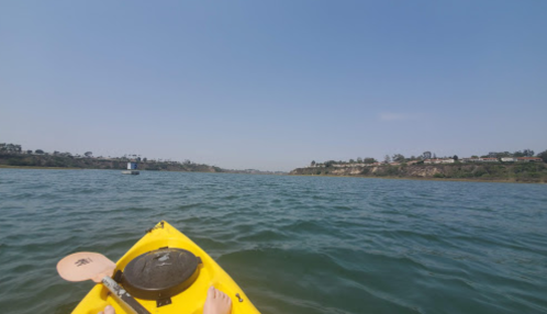 A view from a yellow kayak on calm water, with a distant shoreline and clear blue sky.