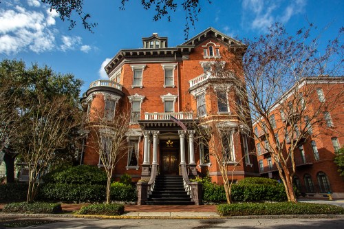 A grand red brick mansion with ornate architecture, surrounded by trees and a clear blue sky.