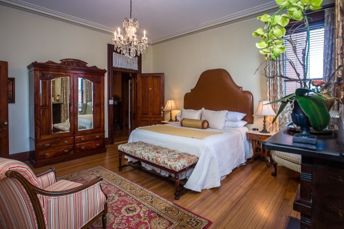 Elegant bedroom featuring a large bed, antique furniture, a chandelier, and decorative plants. Warm, inviting atmosphere.