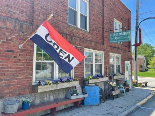 A brick building with a flag reading "OPEN" and flower boxes outside, featuring a sign for Kelsea Designs.