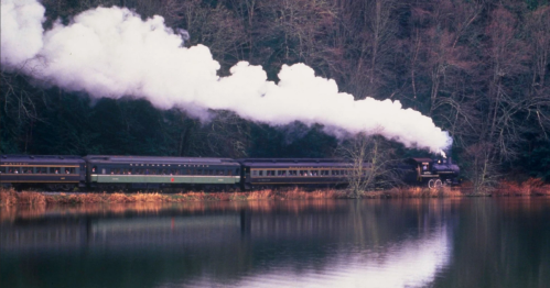 A steam train travels along a river, releasing white smoke against a backdrop of trees and a calm water surface.