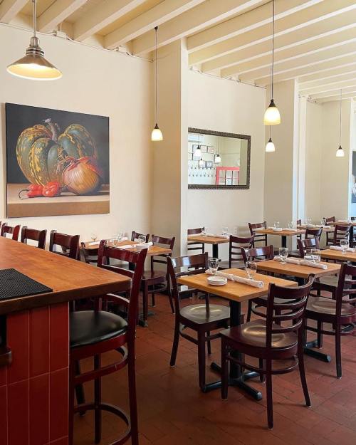 A cozy restaurant interior with wooden tables, chairs, and a large painting of vegetables on the wall.