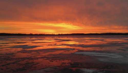 A vibrant sunset casts orange and pink hues over a frozen lake, reflecting the colorful sky on the icy surface.