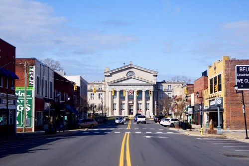 A wide street lined with shops leads to a grand building with columns under a clear blue sky.